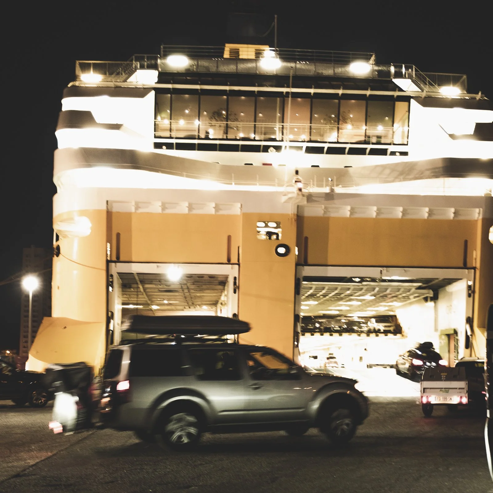 Vue nocturne d'un ferry ou navire de croisière avec plusieurs voitures garées dessous et des passagers sur le pont supérieur, éclairé par des lumières, fond noir.