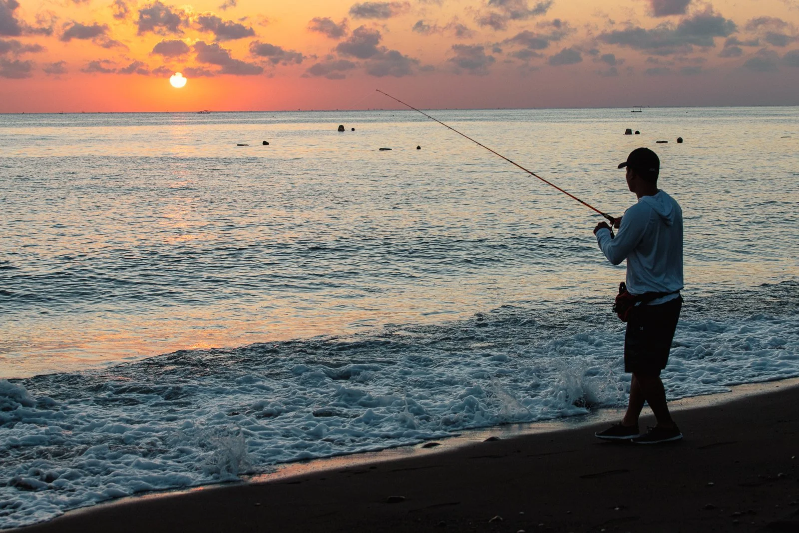 Un homme pêcheur en silhouette au bord de la mer au coucher du soleil, avec le soleil orange dessous des nuages roses et violets, et de petites vagues frappant le rivage.
