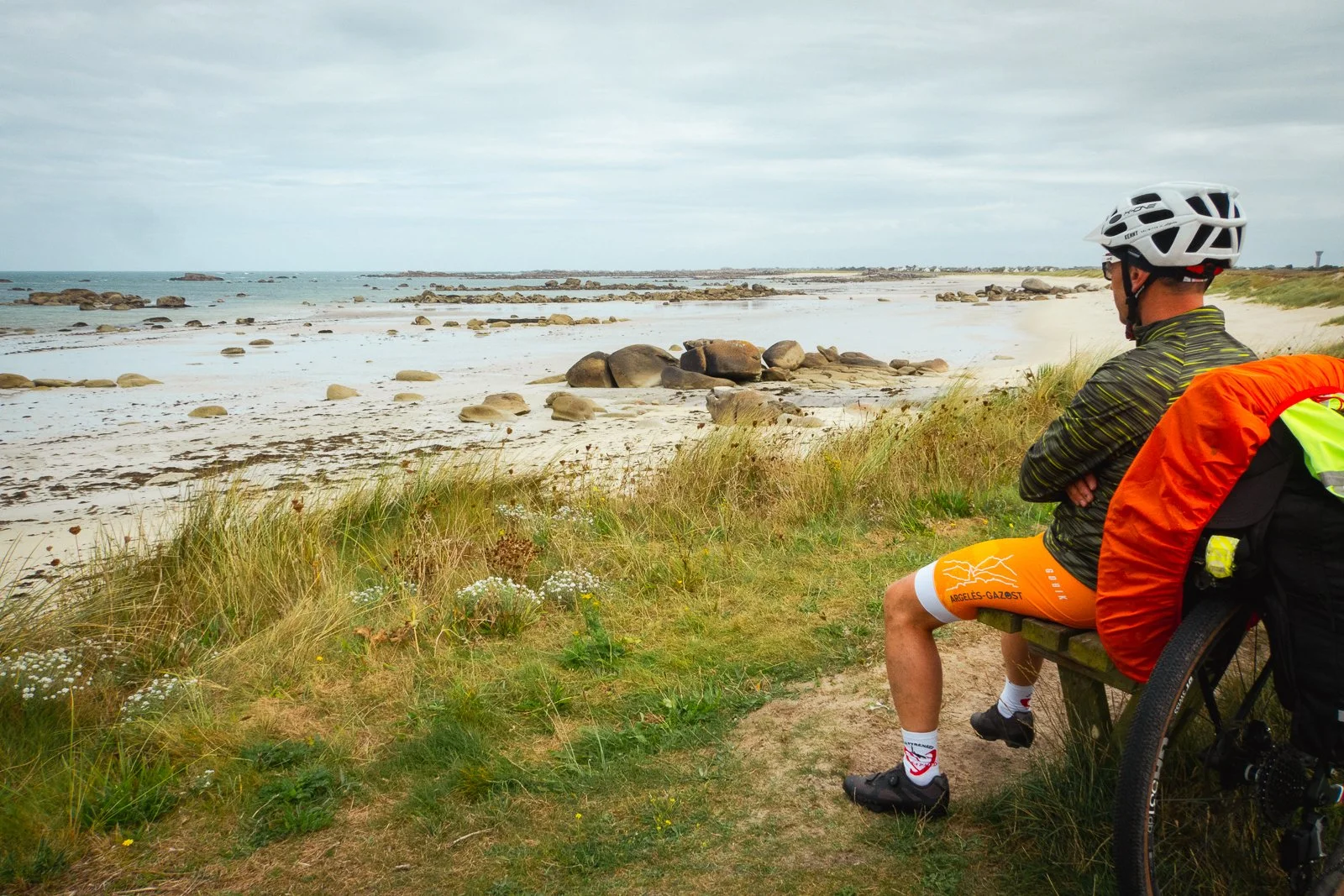 Un homme en vêtements de cyclisme, portant un casque, assis sur un banc près d'une plage avec des rochers, regardant la mer, avec un vélo à côté.