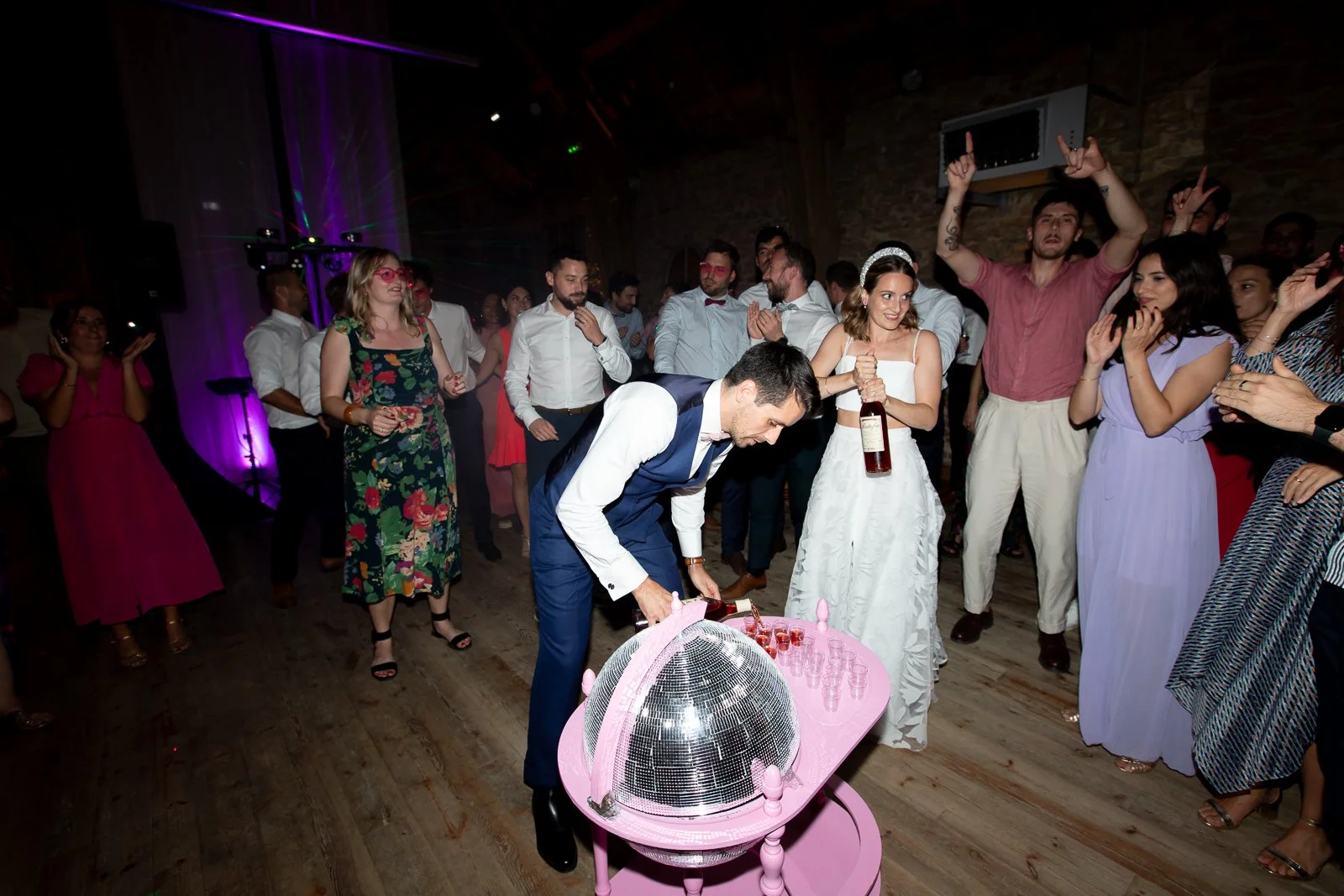 Un groupe de gens danse et s'amuse lors d'un mariage, avec un homme en costume bleu et une femme en robe blanche au centre, entourés de personnes joyeuses portant des vêtements colorés.