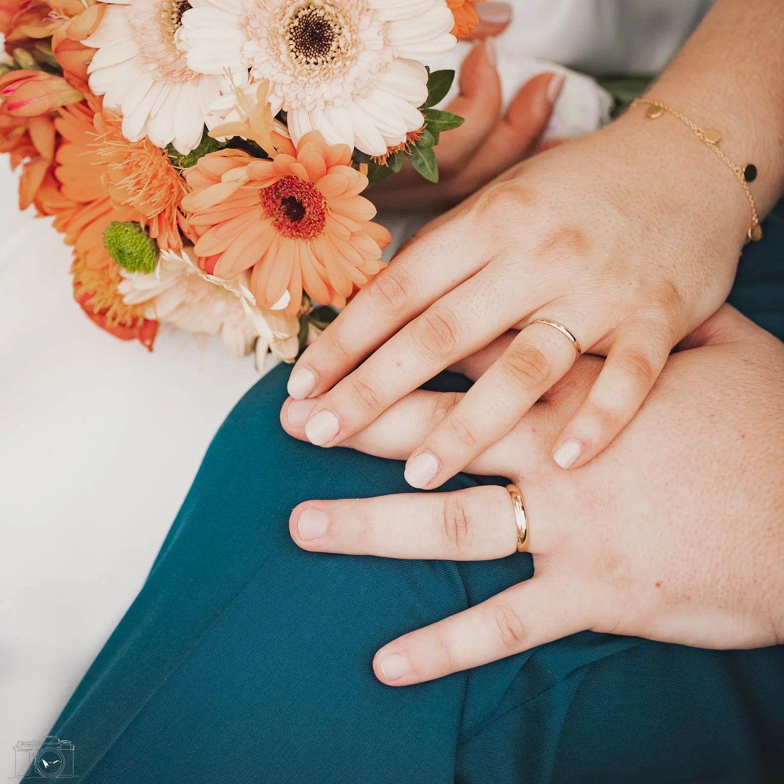 Mains croisées avec bagues, mains tenant un bouquet de fleurs roses, blanches et orange, une personne porte une bracelet doré et une autre une chemise blanche.