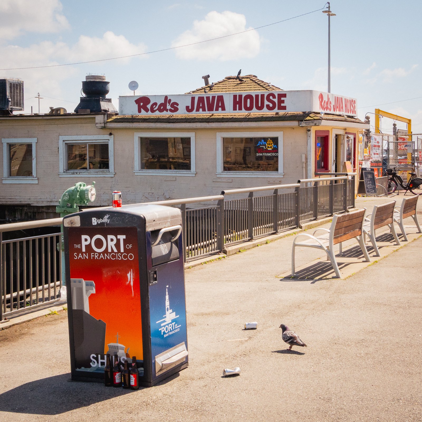 À l'extérieur d'un bâtiment avec un panneau indiquant 'Red's JAVA HOUSE', il y a une enseigne lumineuse avec la silhouette de San Francisco, en face d'une poubelle de recyclage portant une affiche 'PORT OF SAN FRANCISCO'. Sur le sol, deux pigeons, un
