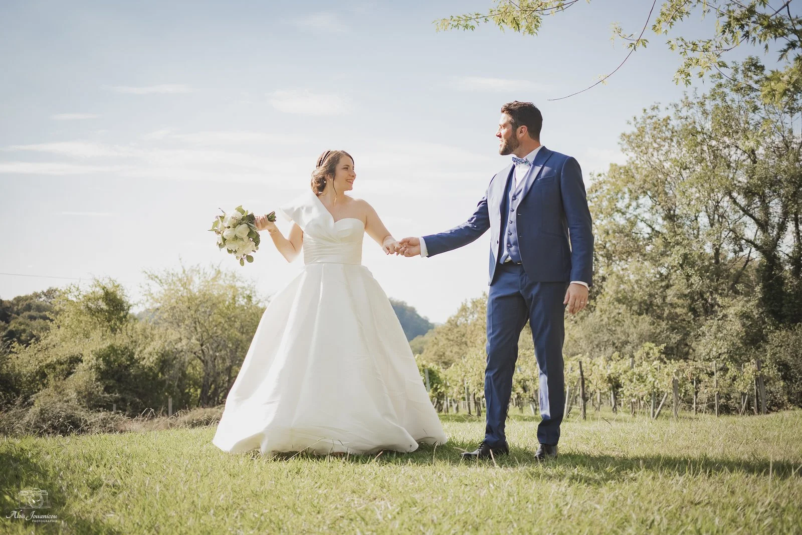 Un couple de mariés, une femme en robe de mariage blanche et un homme en costume bleu, se tenant la main dans un champ avec des arbres et un ciel en arrière-plan.