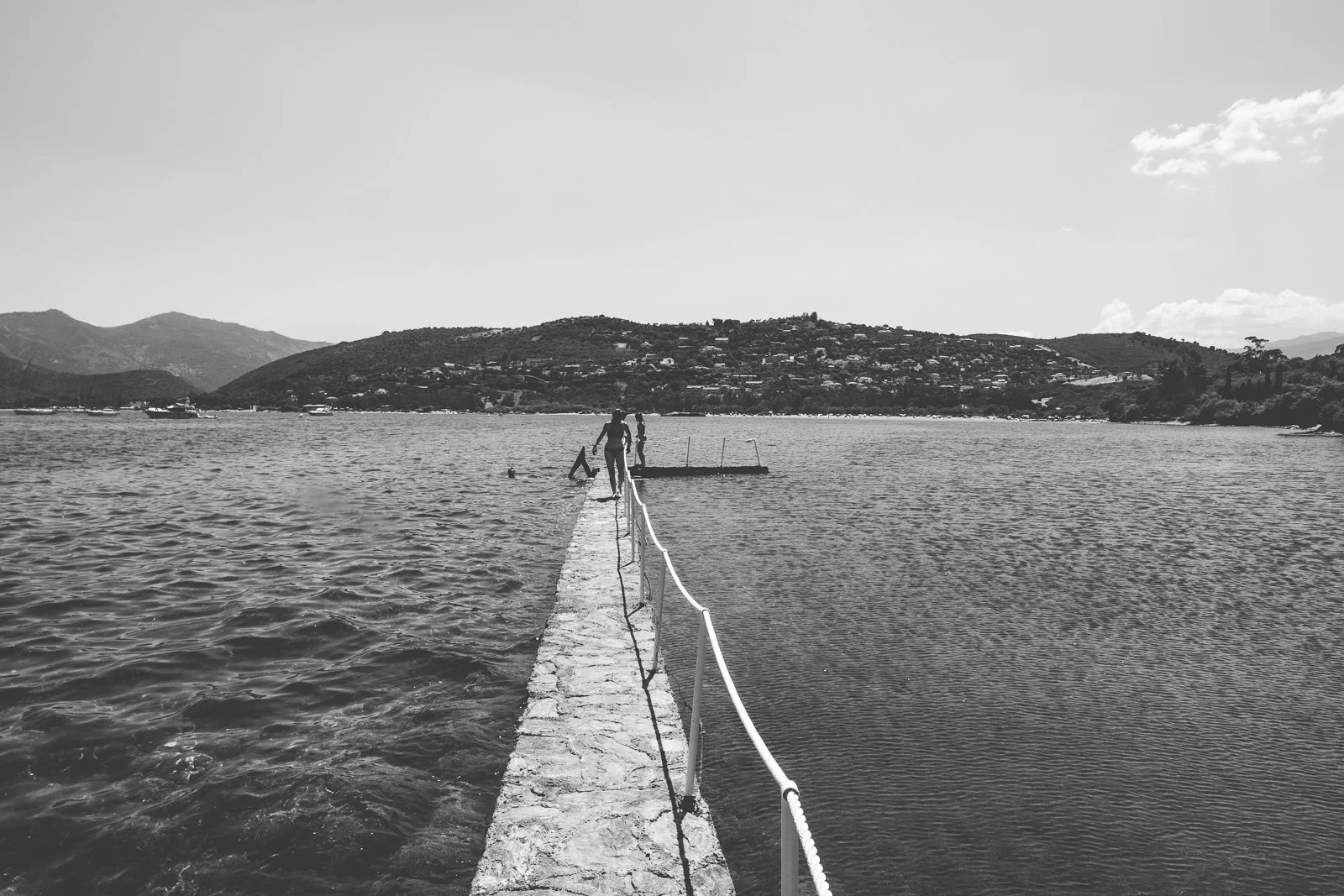 Deux personnes marchent sur une jetée en pierre qui s'étend dans un lac ou une mer, avec des montagnes en arrière-plan, dans un paysage noir et blanc.