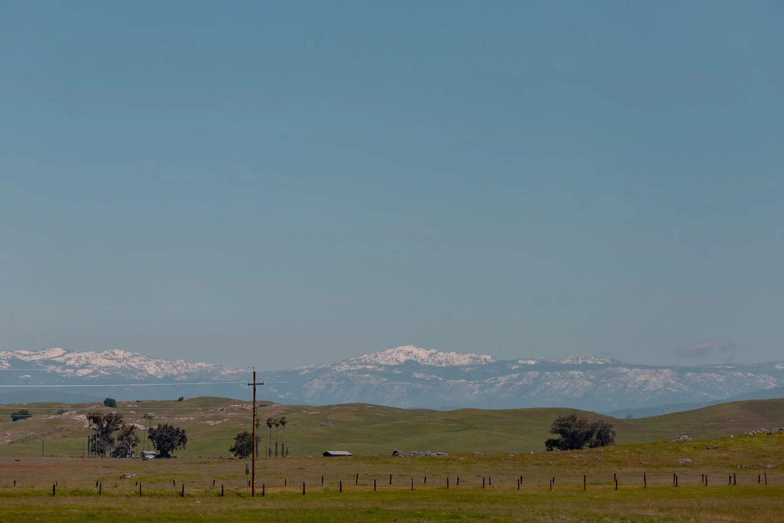 Paysage avec collines vertes, arbres, ligne électrique, et montagnes enneigées au loin sous un ciel clair.