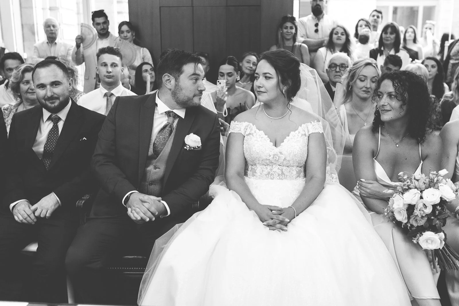 Une photo en noir et blanc d'un mariage avec le couple principal assis au centre, entouré de témoins et invités lors d'une cérémonie en intérieur.
