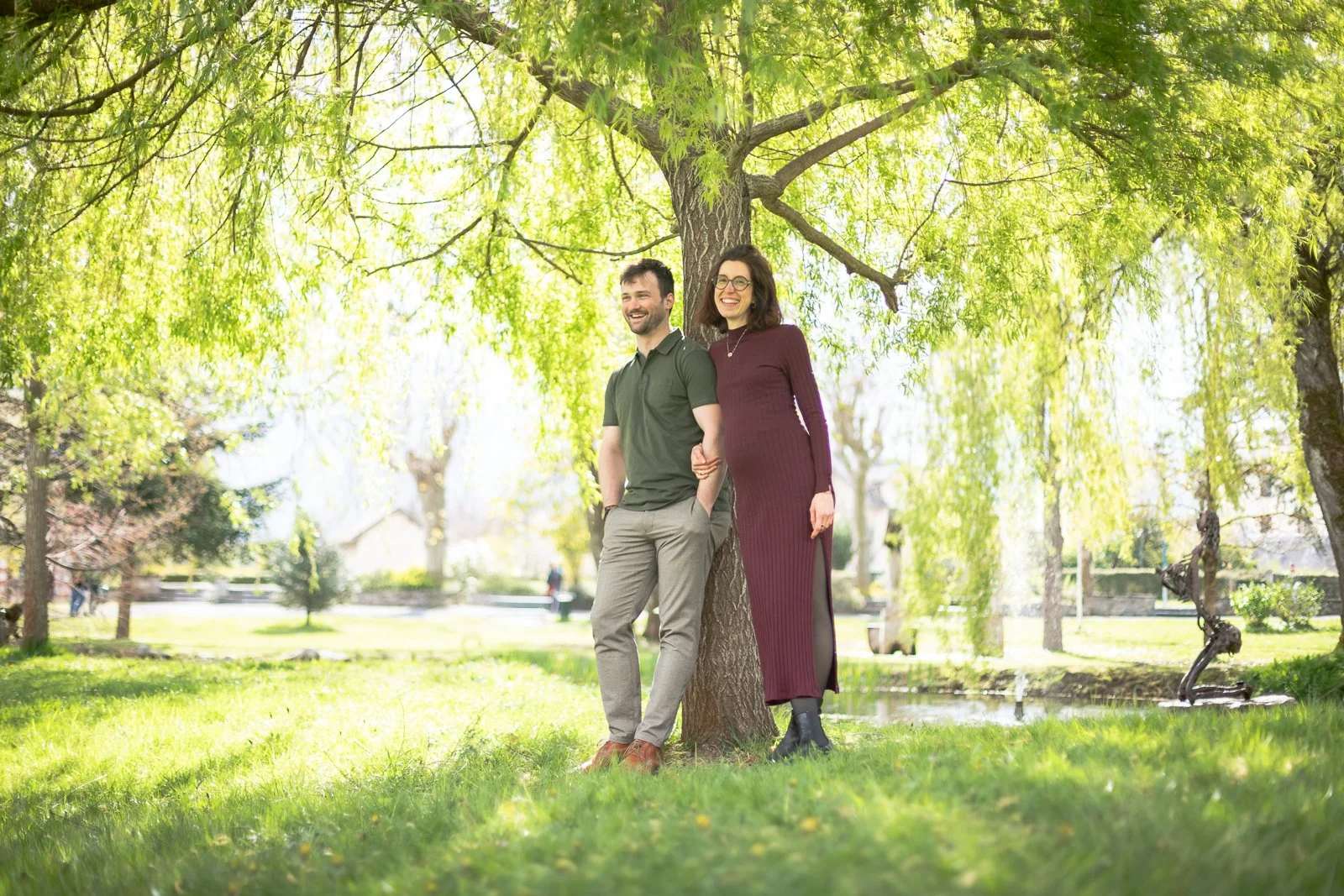 Un homme et une femme souriants, debout sous un arbre dans un parc, pendant une journée ensoleillée.