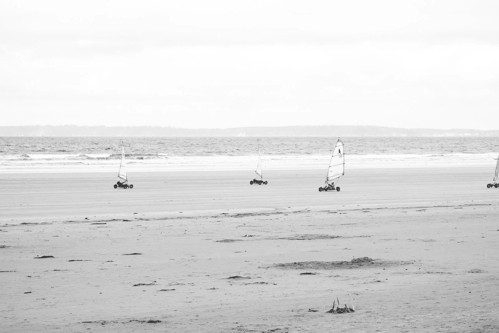 Trois landaus de plage avec des voiles sont alignés sur la plage, près de la mer. L'arrière-plan montre l'océan et un ciel nuageux.