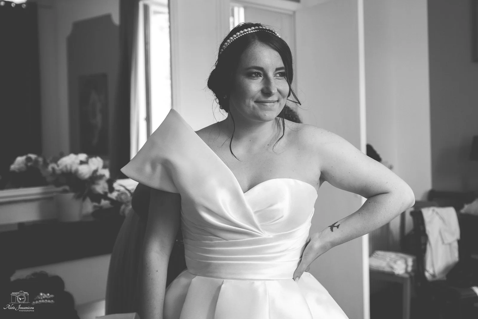 Une femme portant une robe de mariée blanche, souriant, dans une pièce avec des fenêtres et des fleurs en arrière-plan.