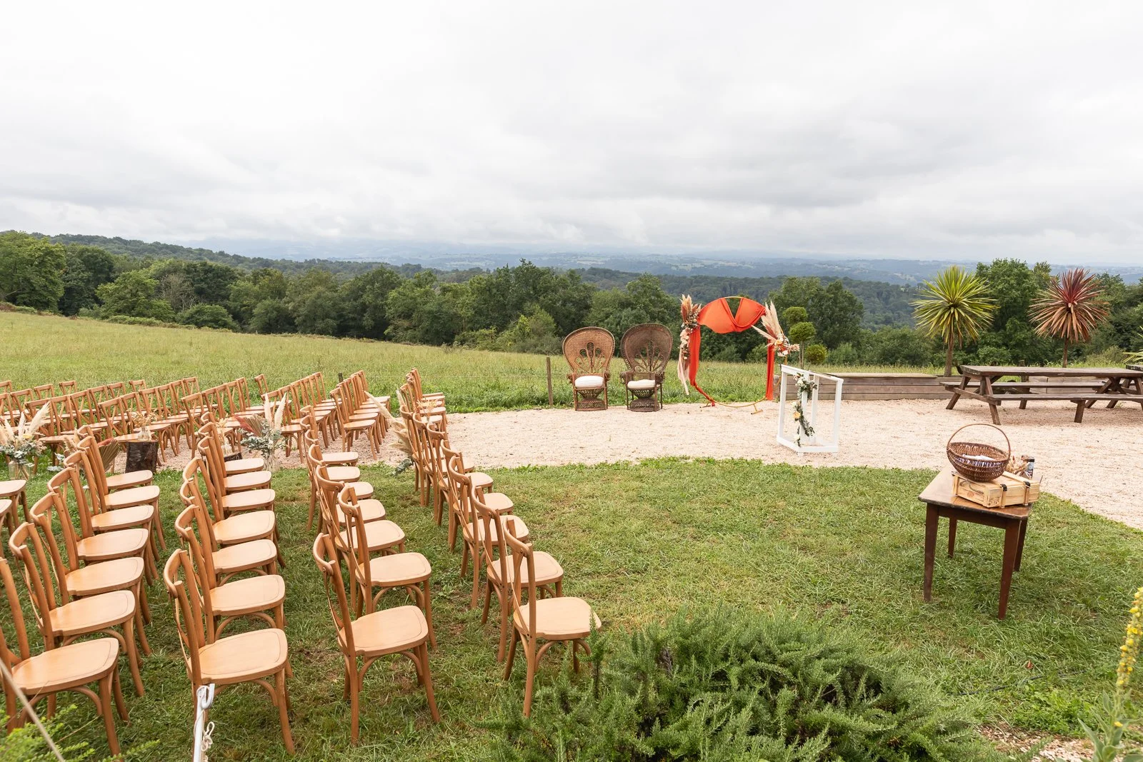 Espace en plein air aménagé pour un mariage, avec des chaises en bois alignées, une arche décorée, deux grands fauteuils, un banc en bois, une table avec un panier et des décorations florales, entouré de verdure et d'un paysage vallonné.