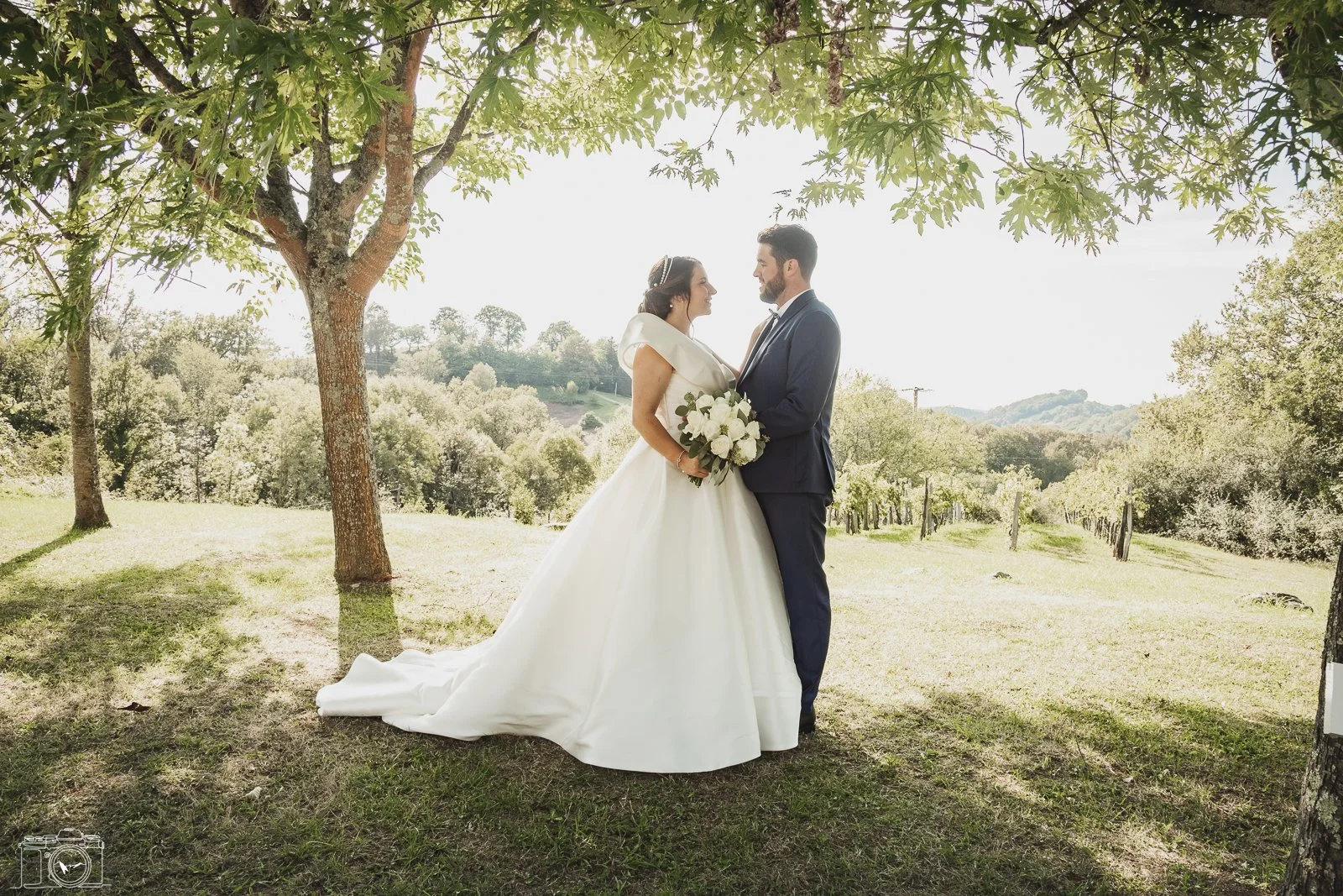 Un couple de mariés se tenant par la main devant un arbre lors d'une séance photo de mariage en plein air, avec un paysage rural en arrière-plan.
