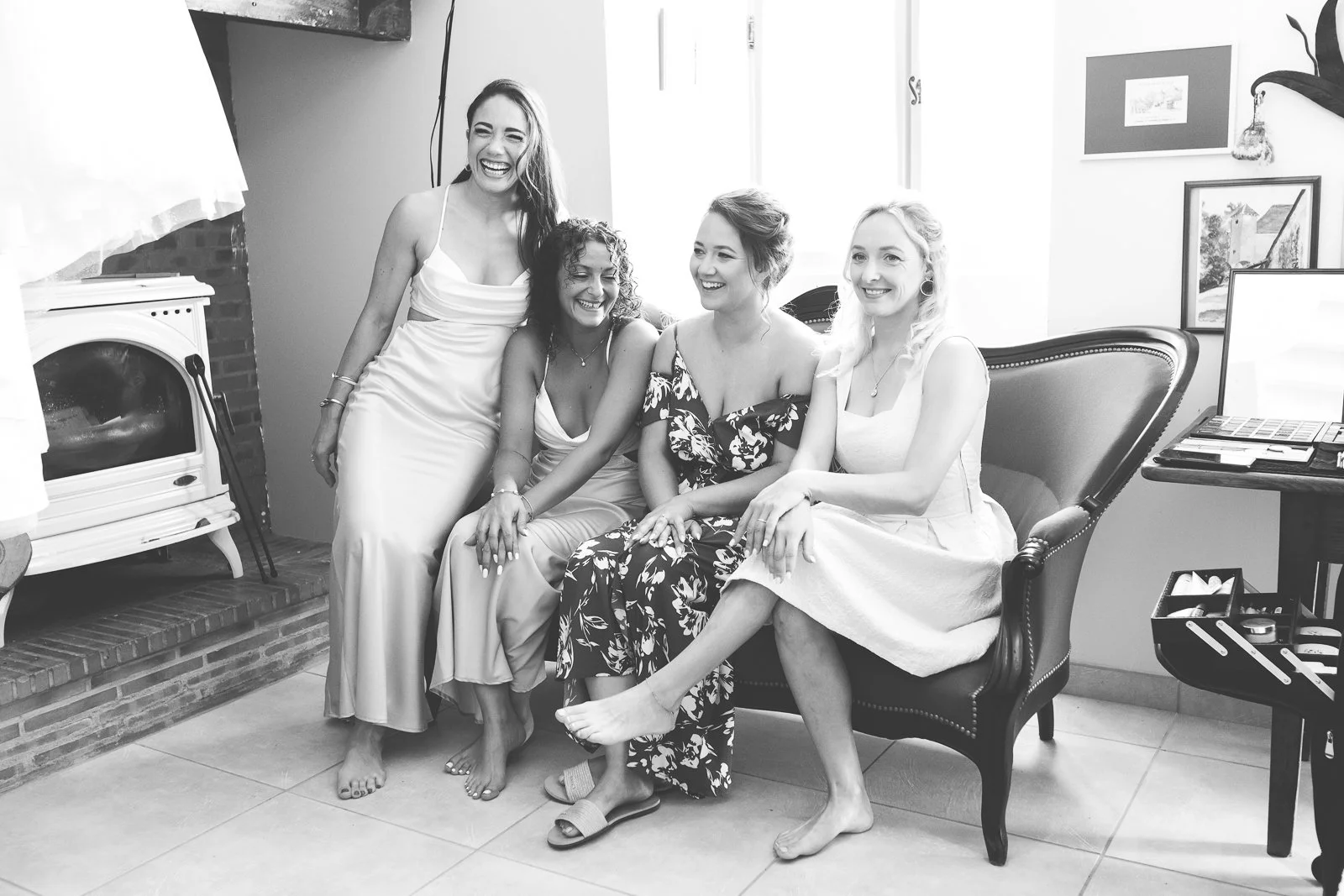 Cinq femmes habillées en vêtements d'été, assises et debout, souriantes, dans un intérieur avec cheminée. Leurs poses et expressions montrent qu'elles sont amies et passent un bon moment ensemble.