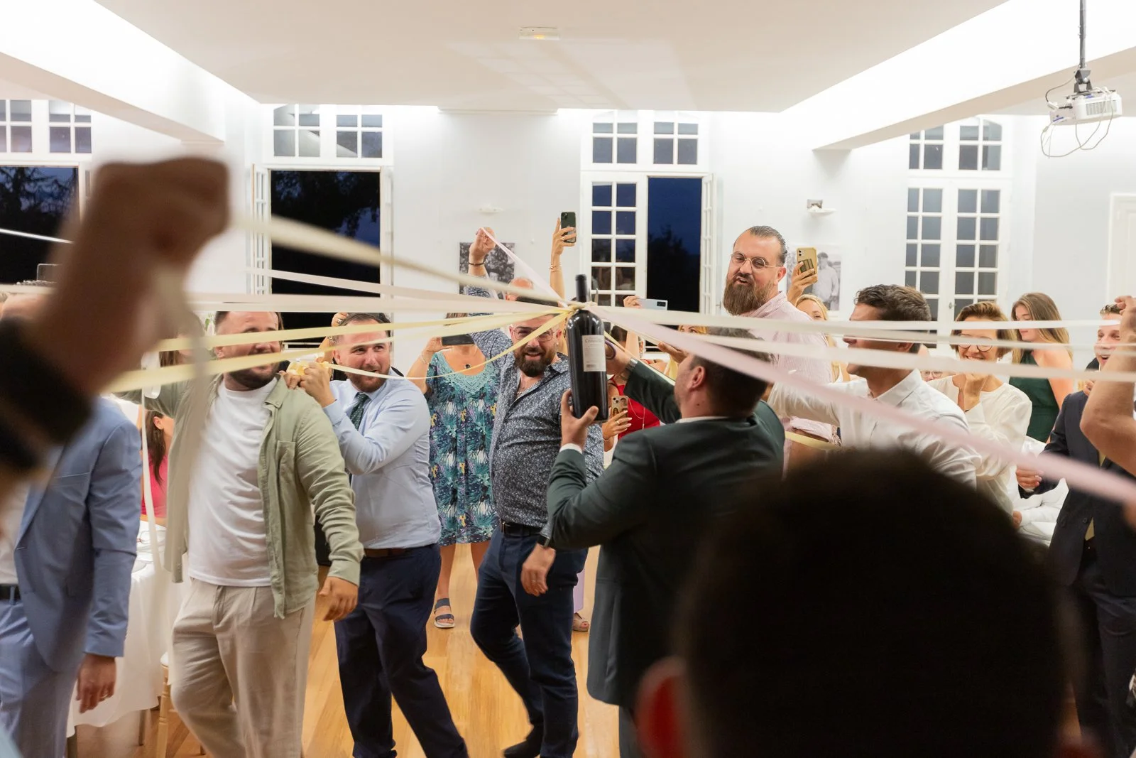 Un groupe de personnes souriantes et joyeuses à une fête ou un mariage, engagées dans une activité ludique avec des rubans tendus d'un côté à l'autre, dans une salle bien éclairée avec de grandes fenêtres et un plafond haut.