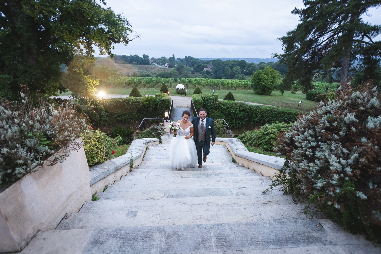 Un couple de mariés descend une allée en pierre dans un jardin verdoyant lors d'un mariage en plein air, avec des arbres et un paysage rural en arrière-plan.