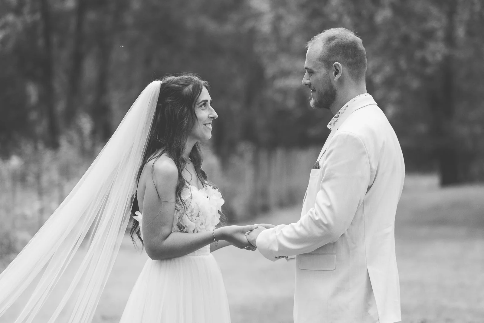 Une mariée et un marié se regardant et se tenant la main lors d'un mariage en plein air. La mariée porte une robe blanche avec un voile, le marié porte un costume blanc. Arrière-plan flou de nature.