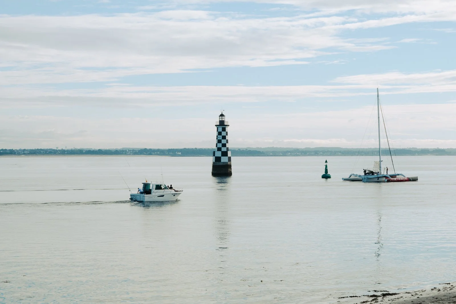 Vue d'un bateau à moteur sur l'eau avec un phare en forme de tour à damier noir et blanc, et un voilier à voile à droite, dans un paysage marin calme sous un ciel partiellement nuageux.