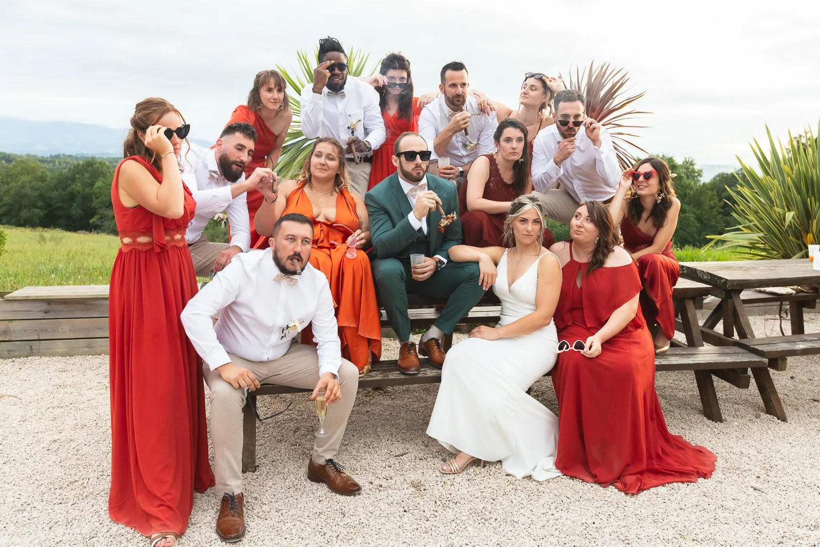 Groupe de personnes à une fête en plein air, certaines portant des robes rouges, un couple assis en tenue de mariage, autour de tables en bois et d'une pelouse verte, avec un ciel nuageux.