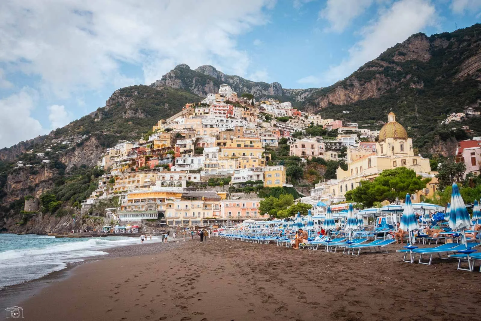 Plage avec parasols bleus, bains de soleil et vue sur des maisons colorées perchées sur une montagne à Positano, Italie.