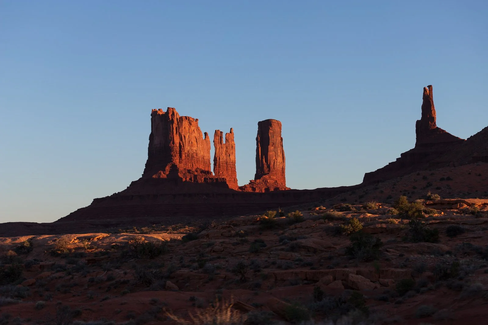Formation rocheuse rouge dans un désert au coucher du soleil, paysage aride avec quelques arbustes, monument naturel en terre battue, silhouettes rocheuses distinctes.