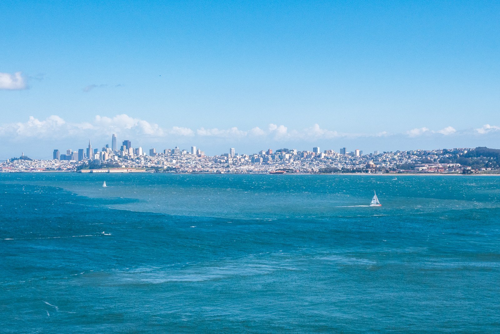 Vue de la baie de San Francisco avec la skyline en arrière-plan et des bateaux à voile sur l'eau bleue.