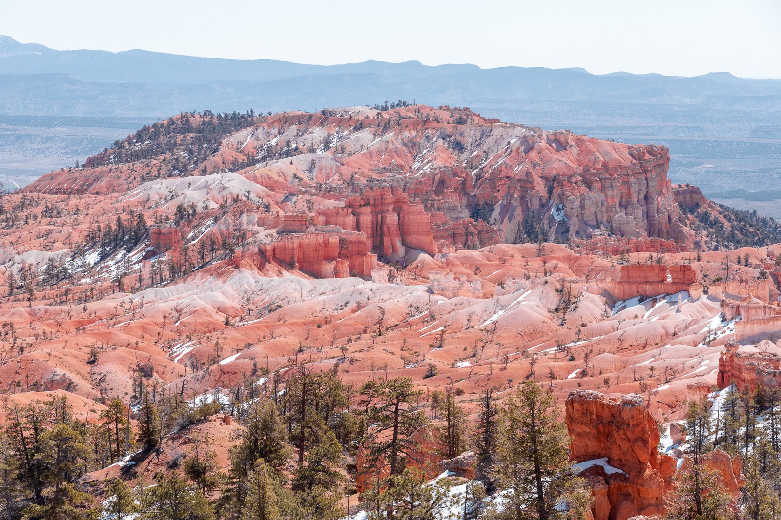 Paysage de montagnes colorées avec des formations rocheuses rouges, quelques arbres, et des taches de neige.
