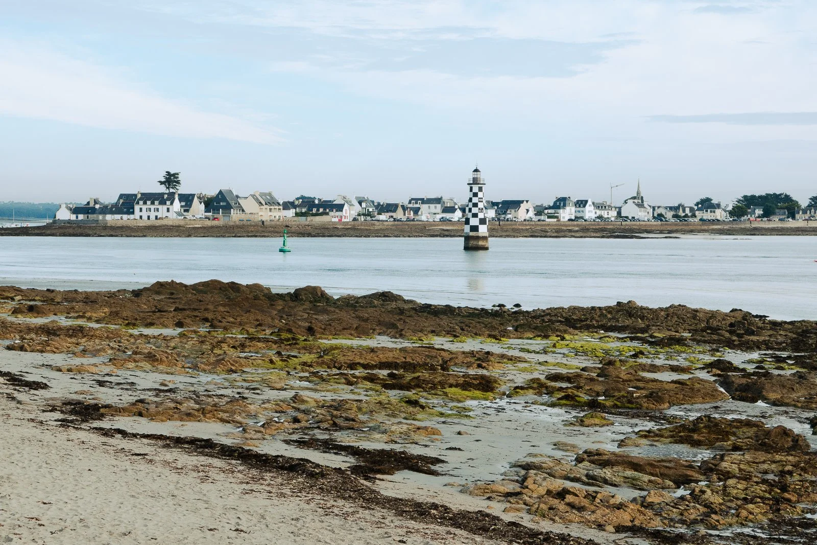 Vue d'une côte avec rochers, eau calme, un phare noir et blanc et un village en arrière-plan avec des maisons blanches et une église.