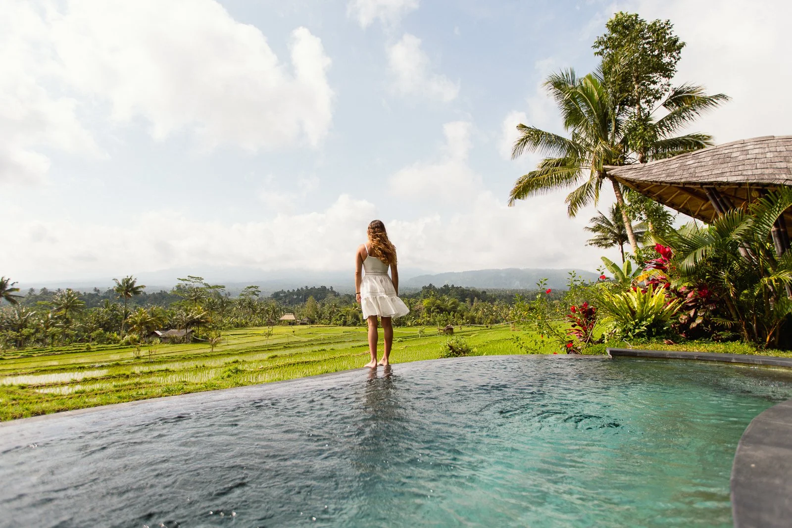 Femme en robe blanche regardant un paysage de rizières vertes avec des palmiers, sous un ciel nuageux, près d'une piscine avec des plantes tropicales.