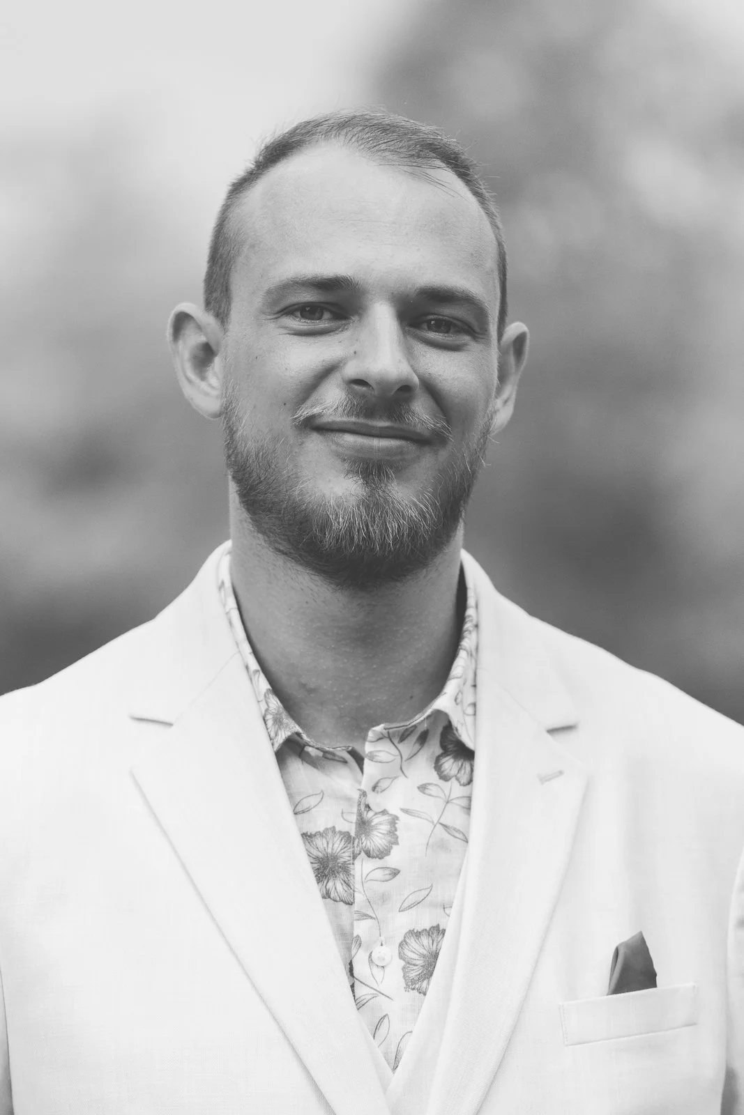 Un portrait noir et blanc d'un homme souriant portant une veste claire et une chemise à motifs floraux, avec un fond flou d'arbres ou de nature.