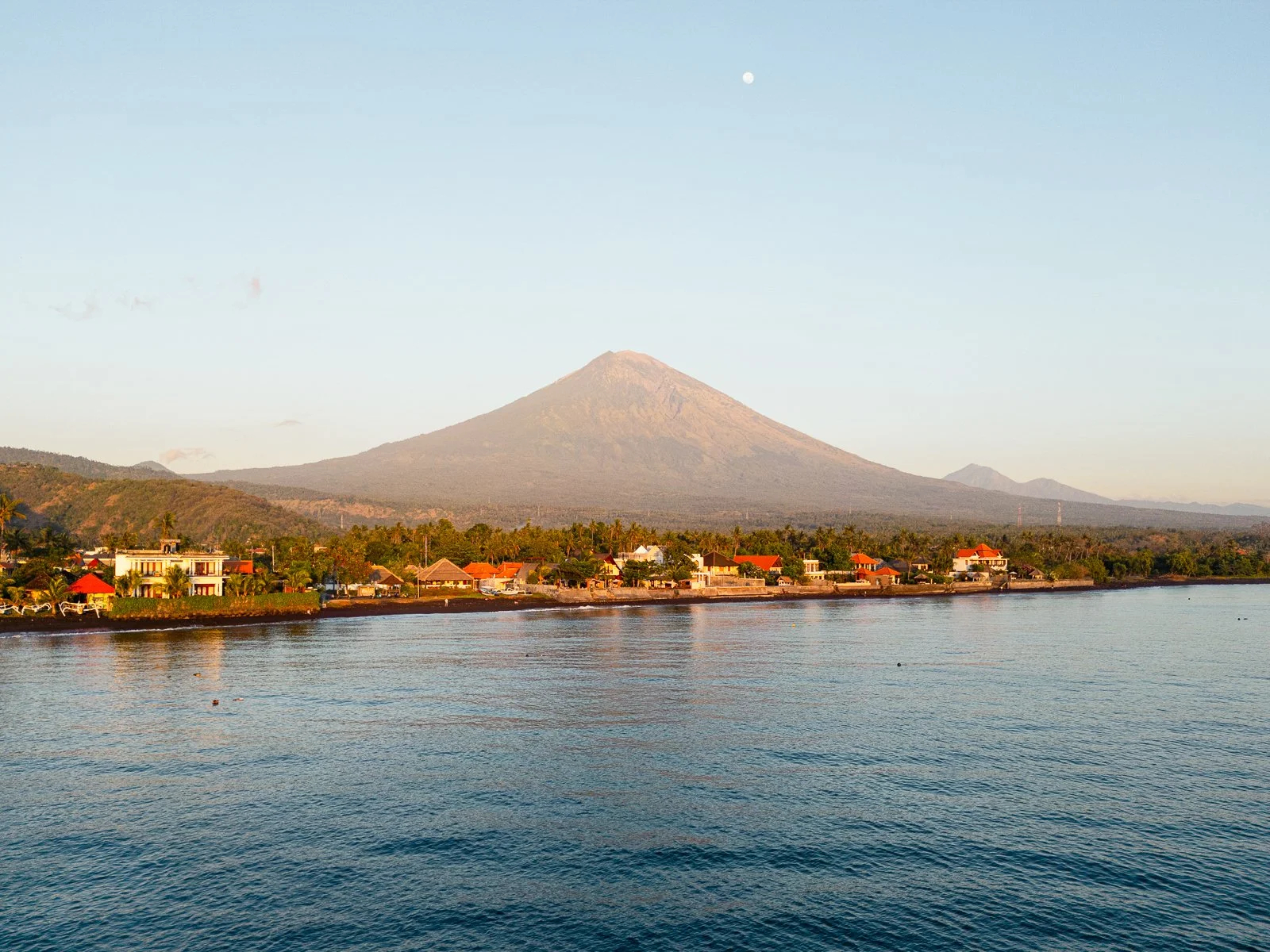 Une vue d'un lac avec des petites maisons en arrière-plan, une montagne imposante avec une crête symétrique, et la lune dans un ciel dégagé.