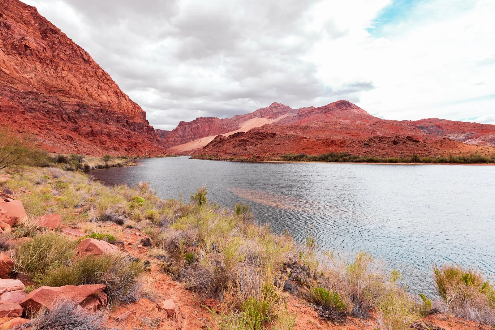 Paysage de canyon avec montagnes rouges et un lac au premier plan, vegetation désertique et ciel nuageux
