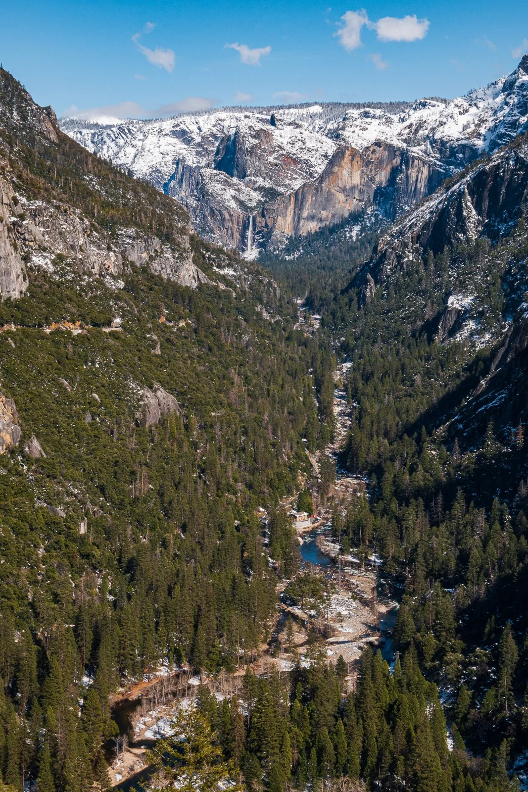 Vue d'une vallée montagneuse avec des sommets enneigés et des forêts de conifères dense.
