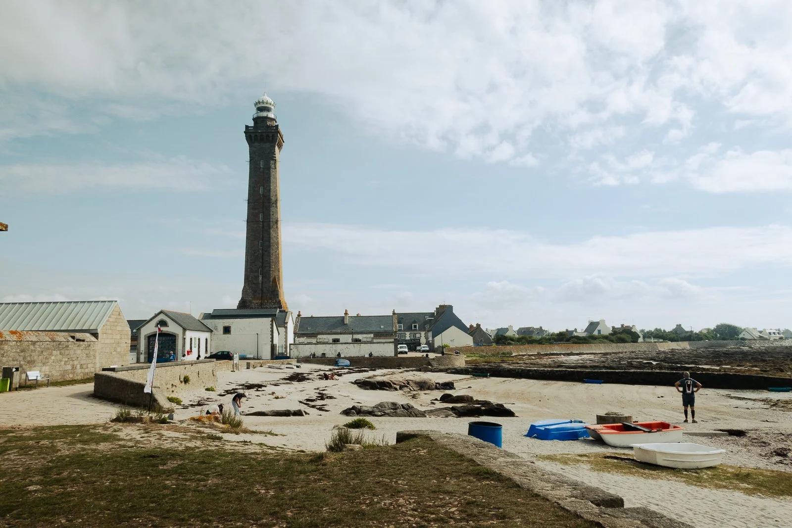 Coucher de soleil sur la plage avec un phare en arrière-plan, des bateaux en bois retournés sur le sable, et une personne regardant la mer.