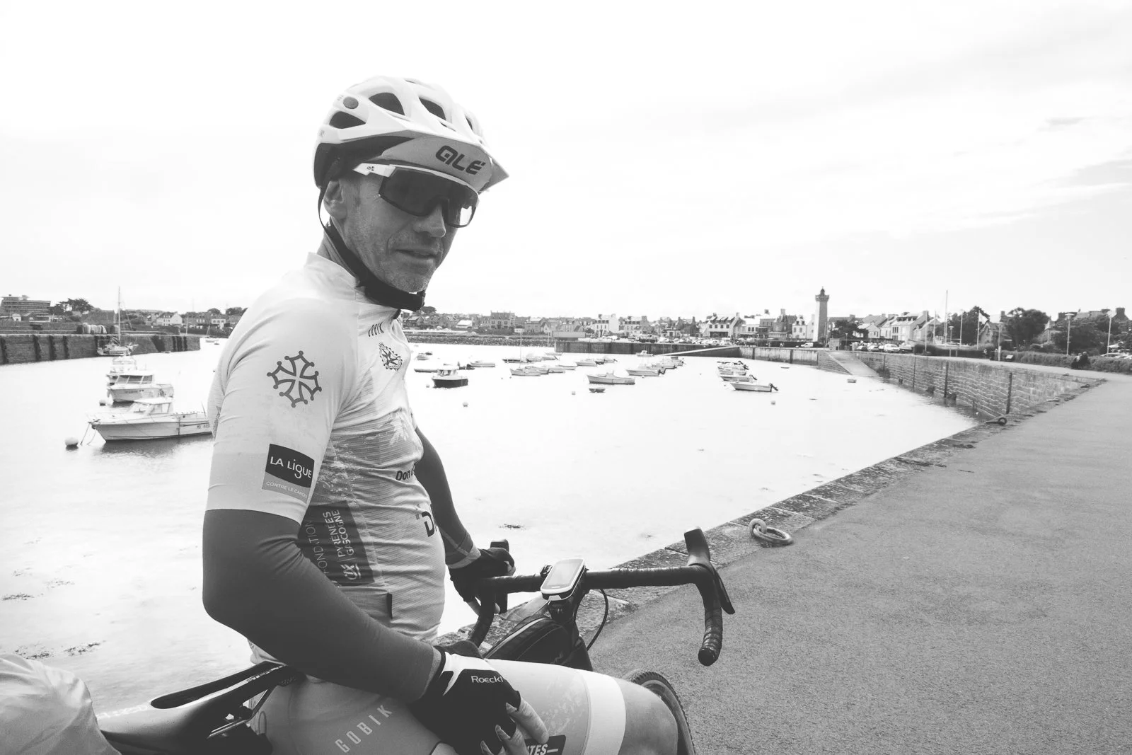 Un cycliste avec équipement de sport, casque et lunettes, tient un vélo près d'un port avec des bateaux, en arrière-plan des maisons et un phare.