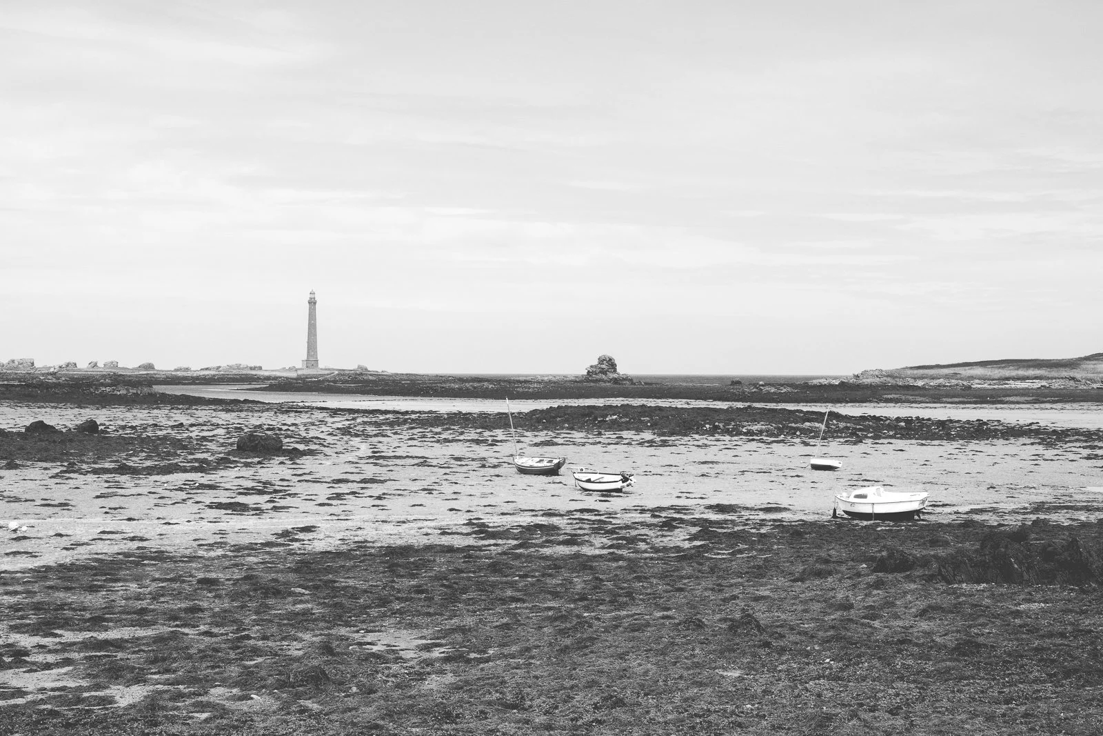 Plage avec trois bateaux échoués, phare en arrière-plan, ciel nuageux, en noir et blanc.