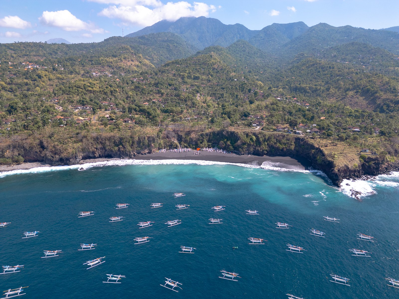 Côte rocheuse avec plage de sable noir et de nombreux bateaux traditionnels en mer, flanquée de montagnes verdoyantes en arrière-plan.