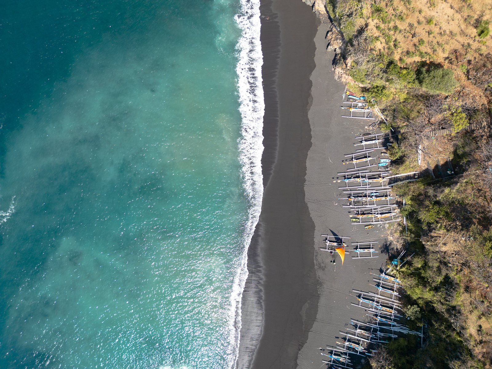 Vue aérienne d'une plage avec des parasols et des voiliers, entourée de végétation et de collines.