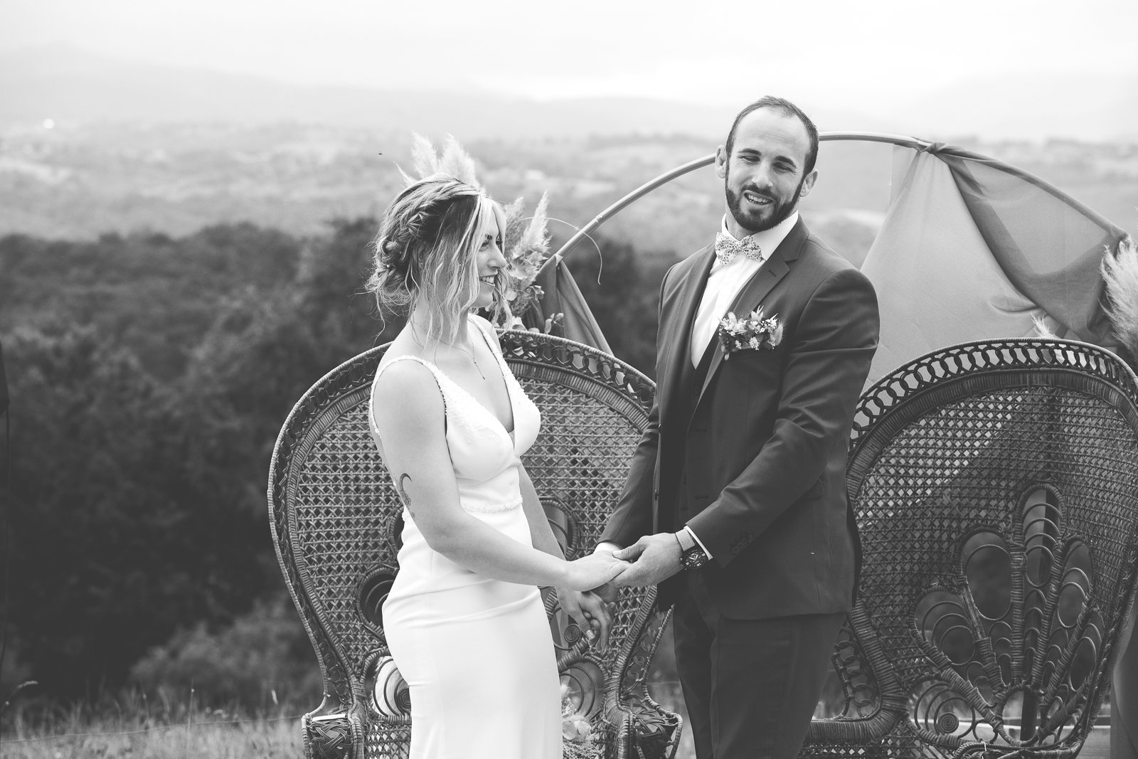 Photographie en noir et blanc d'un mariage, montrant un couple tenant leurs mains et échangeant un regard heureux, avec un paysage naturel en arrière-plan.