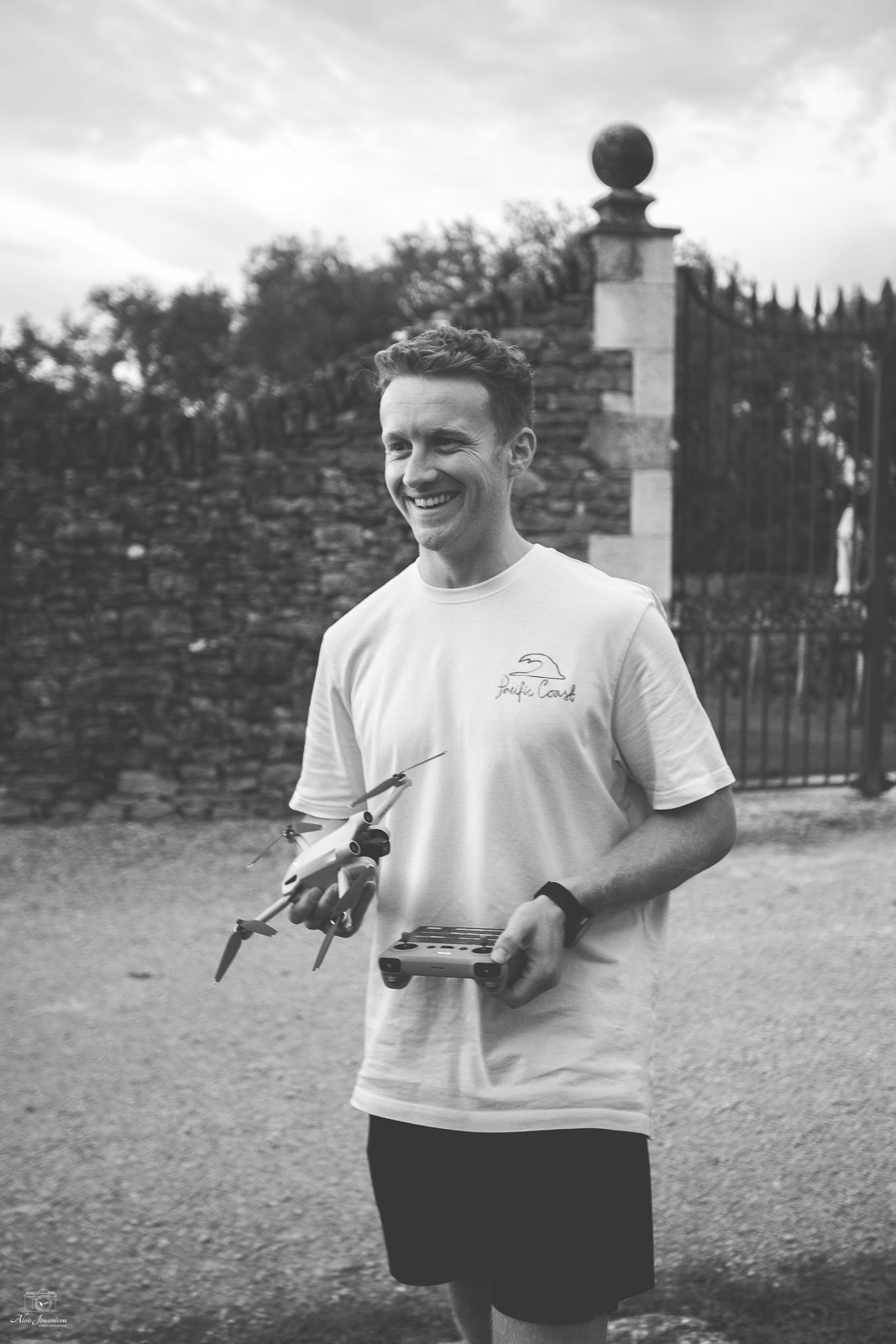 Un homme souriant tenant un drone et une télécommande, en extérieur, en noir et blanc.