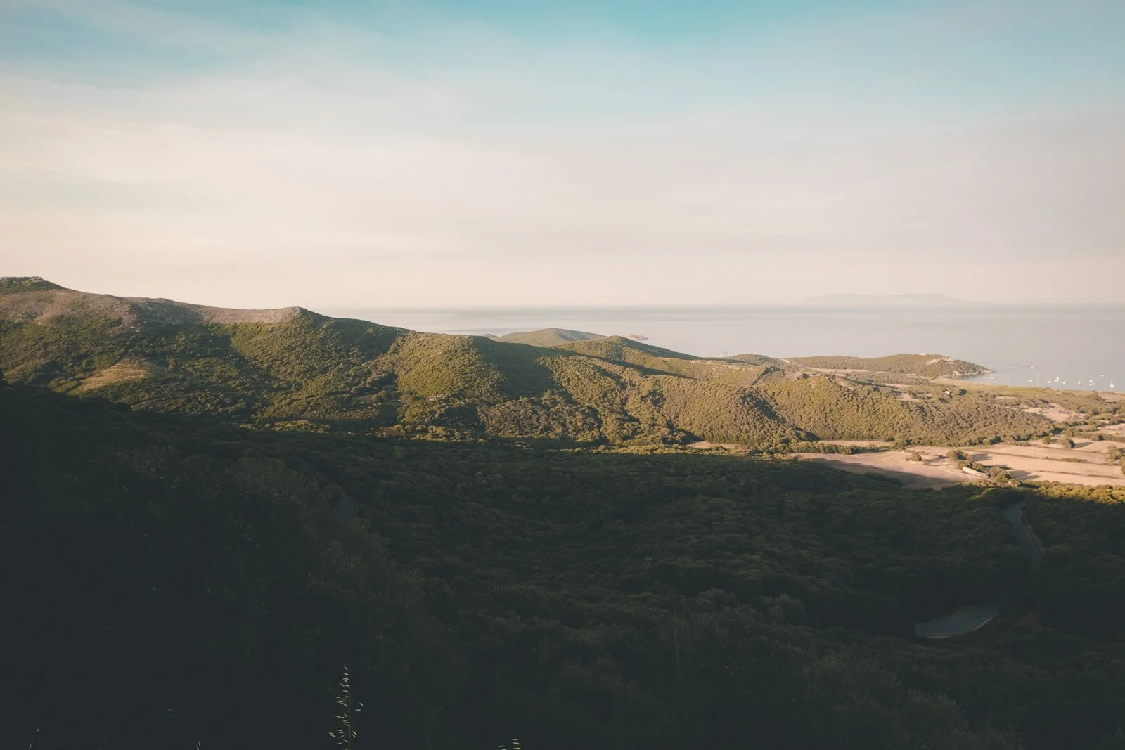 Paysage de collines verdoyantes avec vue sur la mer et quelques voiliers au loin.