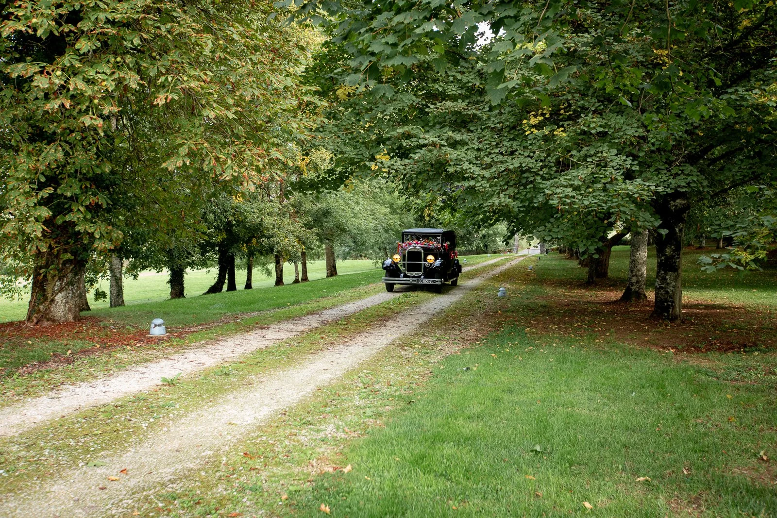 Voiture ancienne noire roulant sur un chemin de gravier dans un parc arboré.