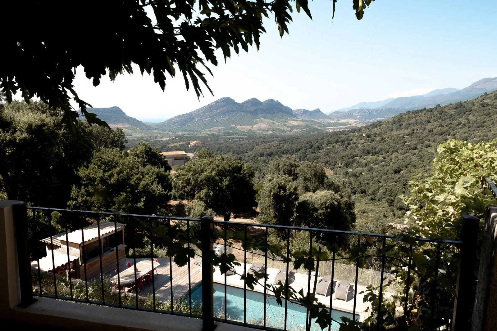 Vue d'un paysage montagneux verdoyant vu depuis une terrasse avec un balcon, des arbres, une piscine, des chaises longues, et des tables en bois, sous un ciel clair.