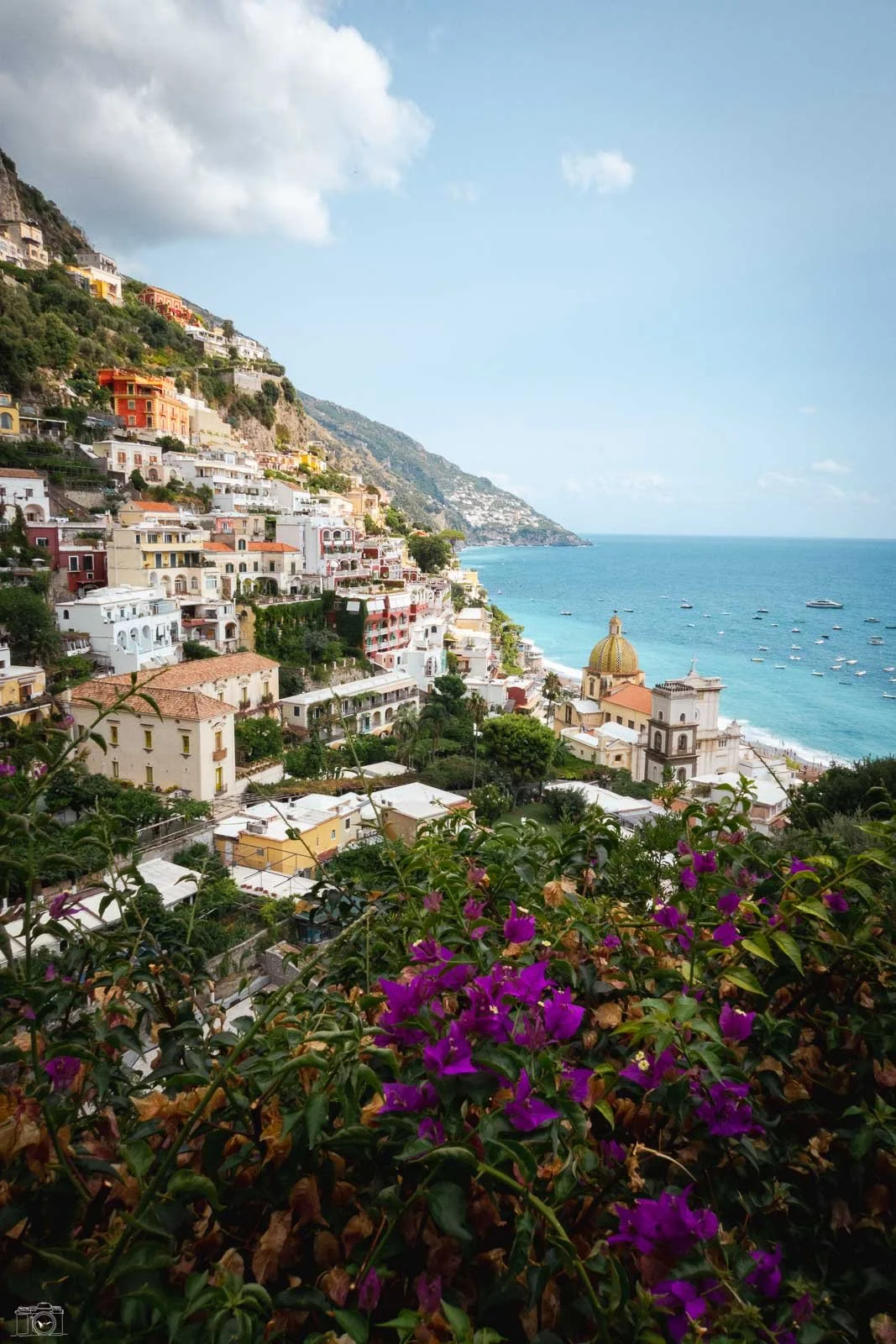 Paysage côtier avec des maisons colorées en escalier sur une colline, vue sur la mer avec des bateaux, arbres et fleurs violettes au premier plan, ciel partiellement nuageux.