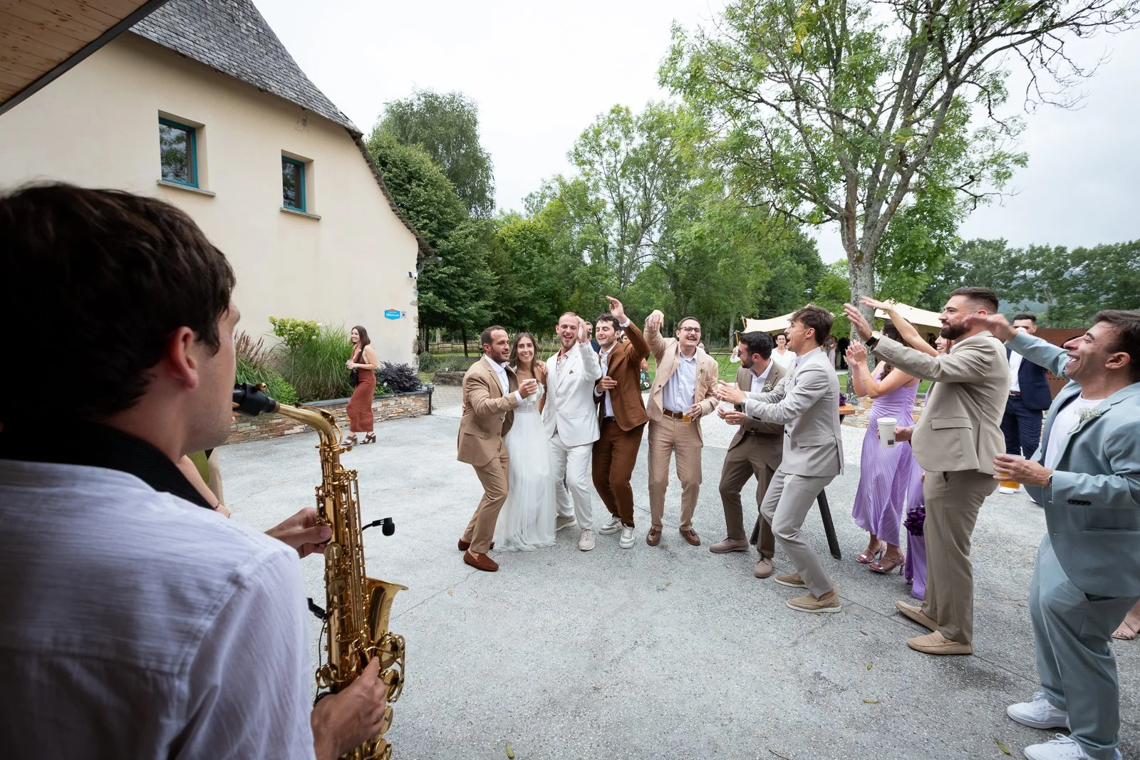 Un groupe de personnes habillées élégamment danse et chante lors d'une fête en extérieur, avec un musicien jouant du saxophone en premier plan.