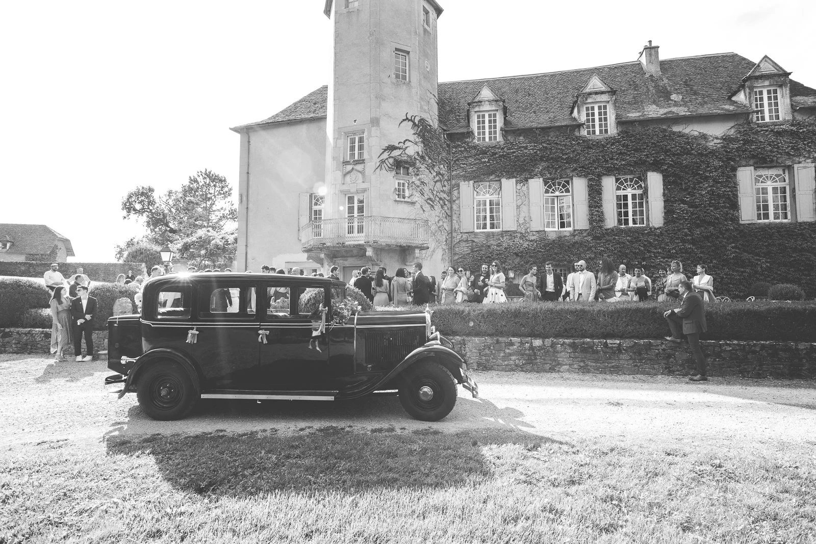 Photographie en noir et blanc d'une assemblée de personnes devant un vieux château avec un garage vintage au premier plan. L'événement semble être une célébration ou un rassemblement social.