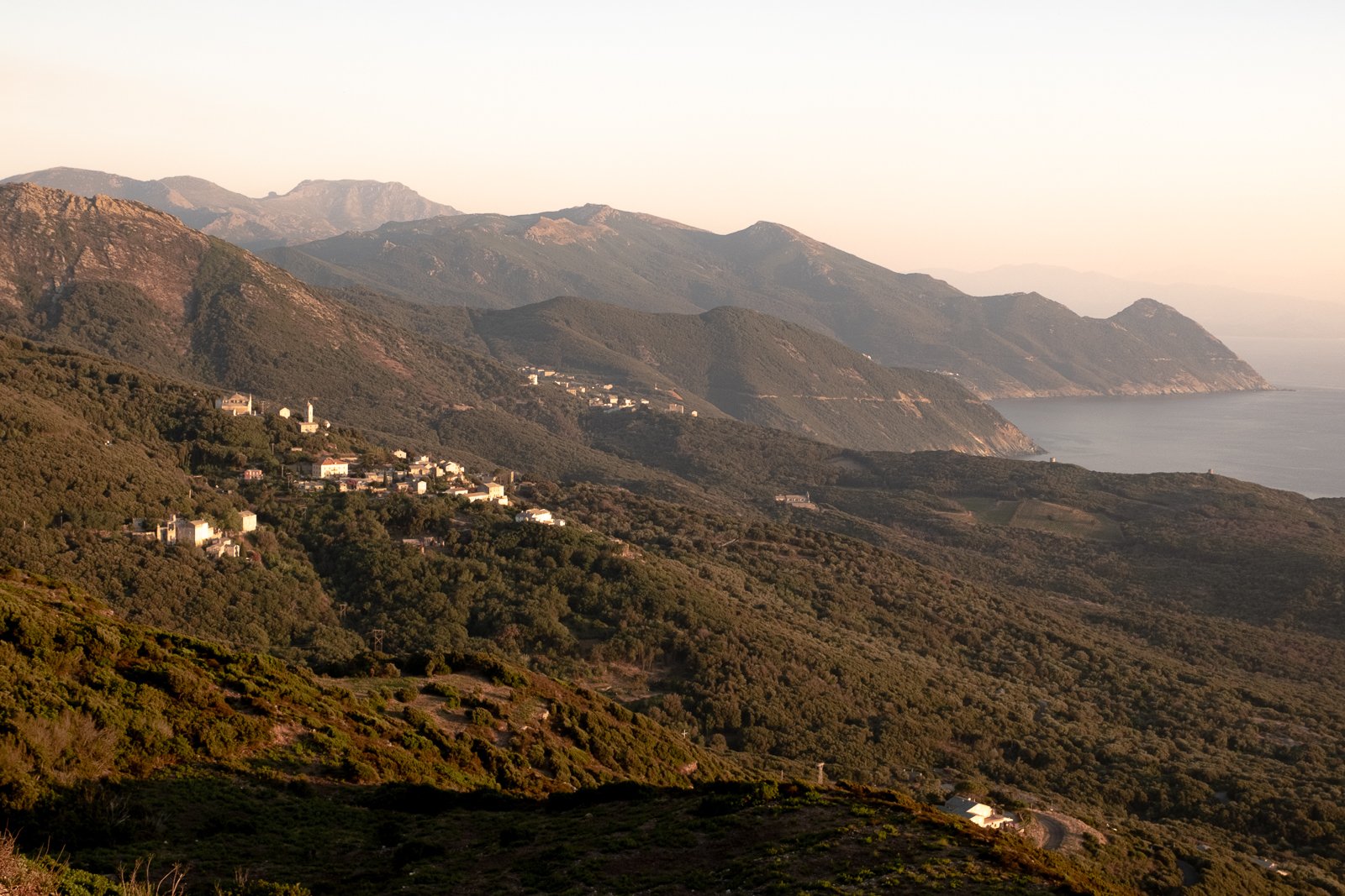 Paysage de montagnes avec des maisons dispersées, vue sur la mer au loin lors d'un coucher de soleil.
