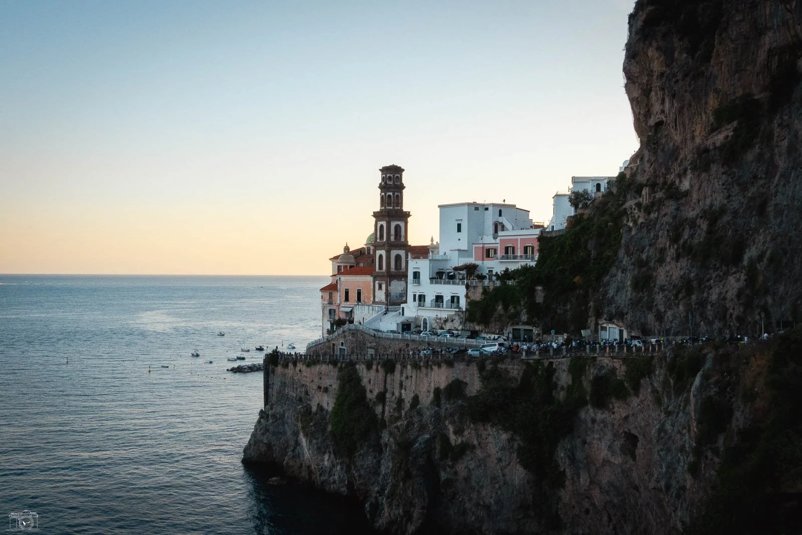 Vue d'une ville côtière construite sur une falaise au coucher du soleil, avec des bâtiments blancs et un clocher en pierre, face à la mer calmement