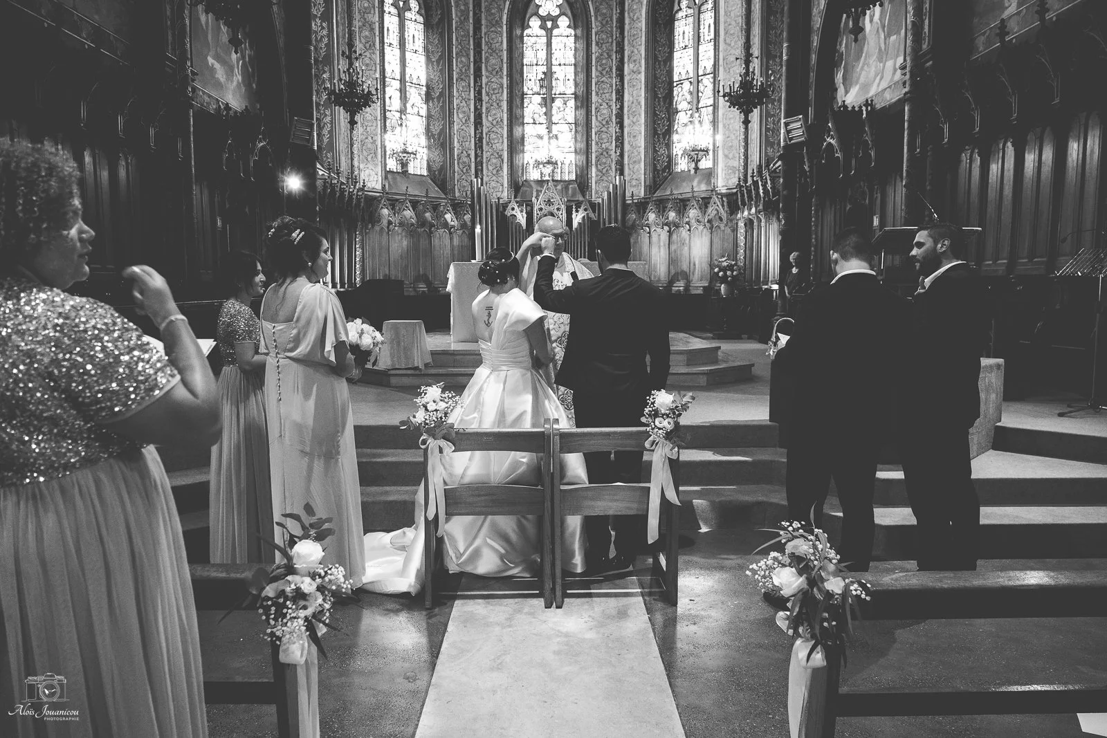 Une cérémonie de mariage religieuse dans une église, avec un couple de mariés, le prêtre et des témoins. Décorations florales et architecture gothique en arrière-plan.