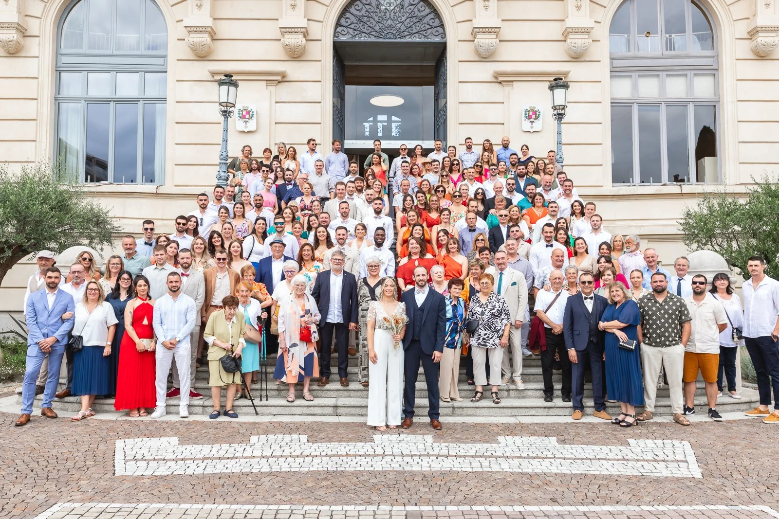 Un grand groupe de personnes posent pour une photo devant un bâtiment historique. Nombreux hommes et femmes, certains en costume ou robe de cérémonie, suggérant un événement festif ou officiel.
