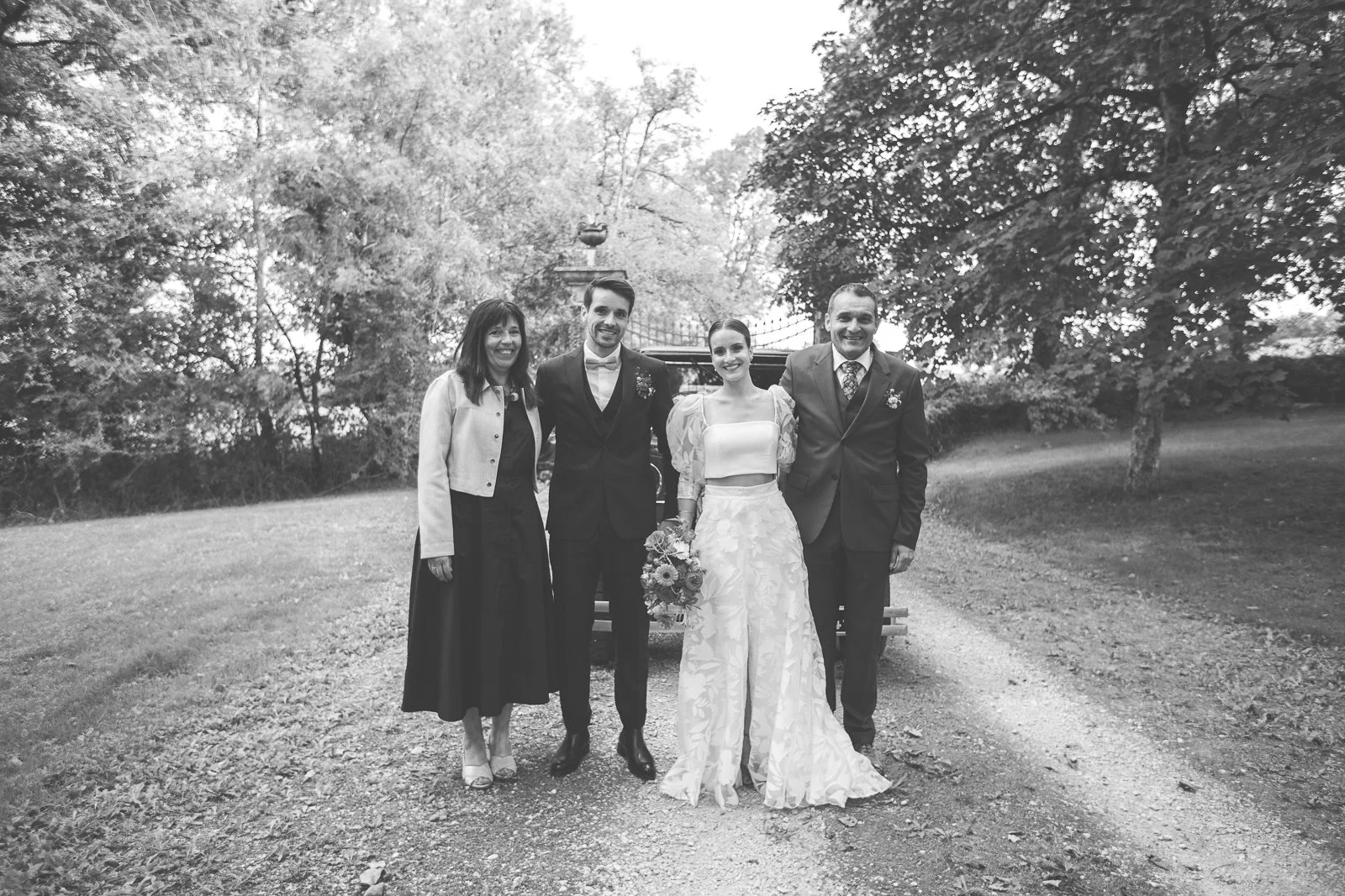 Photo en noir et blanc d'un groupe de cinq personnes lors d'un mariage, posant devant une voiture ancienne dans un parc avec des arbres.