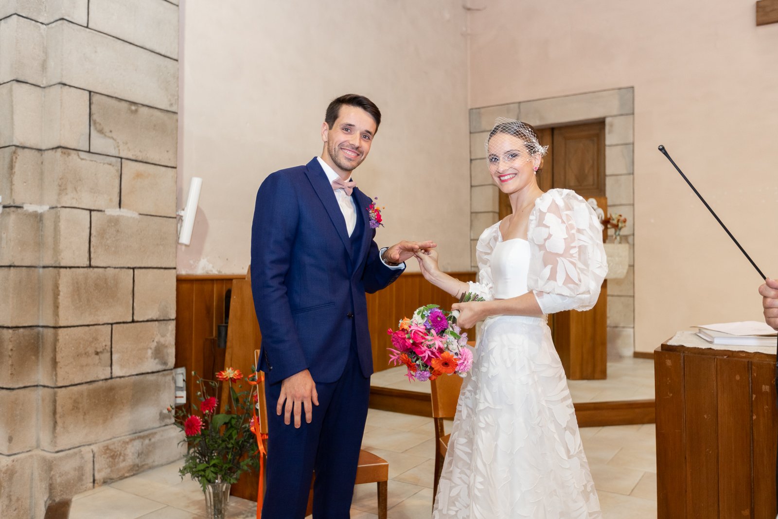 Un couple lors de leur mariage, la mariée en robe blanche avec un bouquet de fleurs colorées, l'homme en costume bleu, dans une salle avec des murs en pierre et une décoration florale.